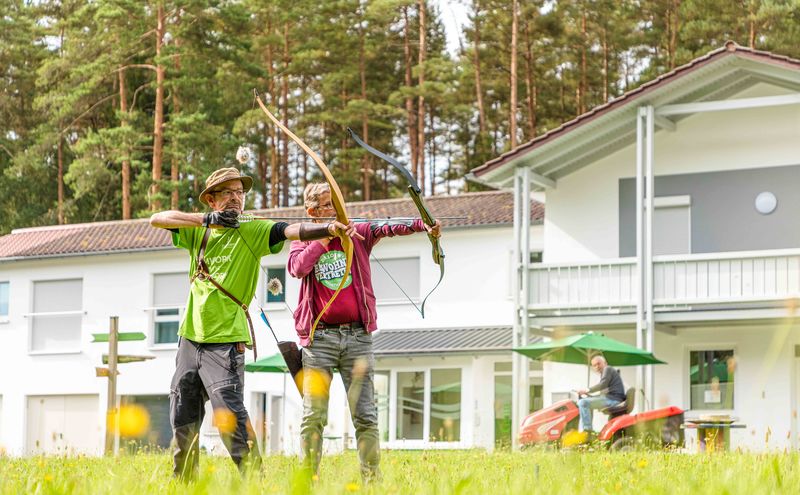 Bogenschießen auf dem Gelände des Grünthaler Hofs Parkstein – Dr. Loew Einrichtung Klient und Mitarbeitender des Grünthaler Hofs Parkstein üben Bogenschießen auf der Wiese vor dem Wohngebäude