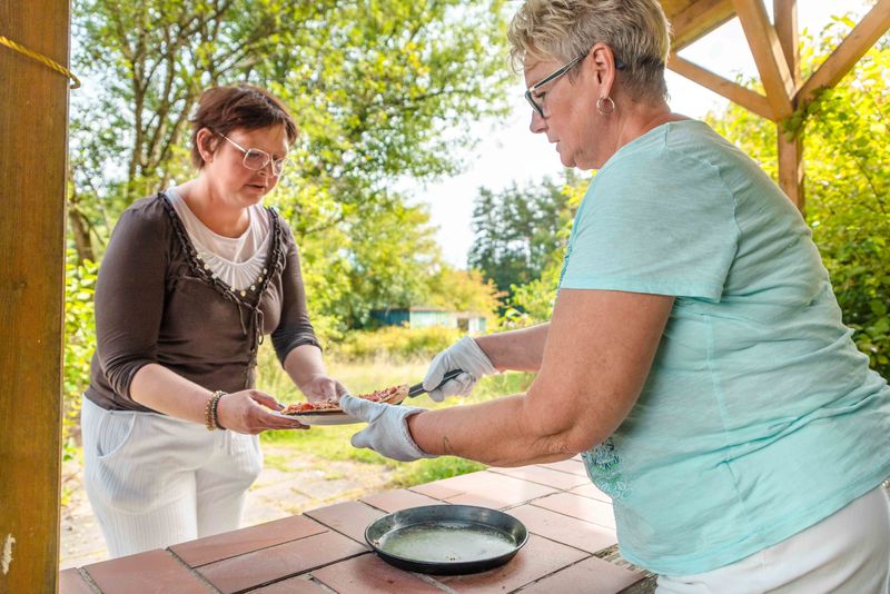 Pizza servieren im Garten – gemeinsames Essen im Grünthaler Hof Parkstein, Dr. Loew Einrichtung Mitarbeitende des Grünthaler Hofs Parkstein reichen einer Klientin frisch gebackene Pizza im Gartenbereich