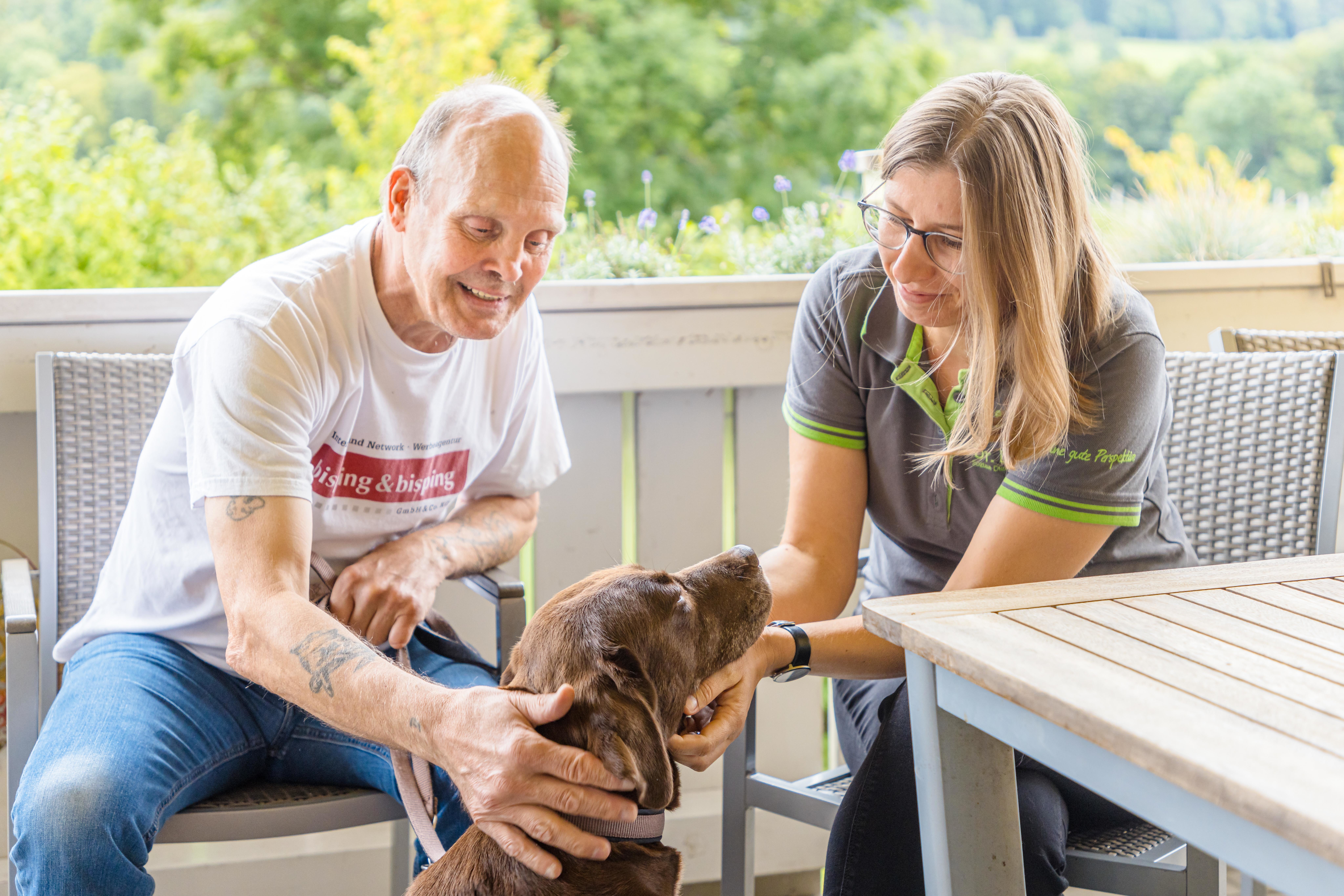 Mann mit geistiger Behinderung in der Einrichtung Dr. Loew gemeinsam mit Betreuerin und Hund auf der Terrasse