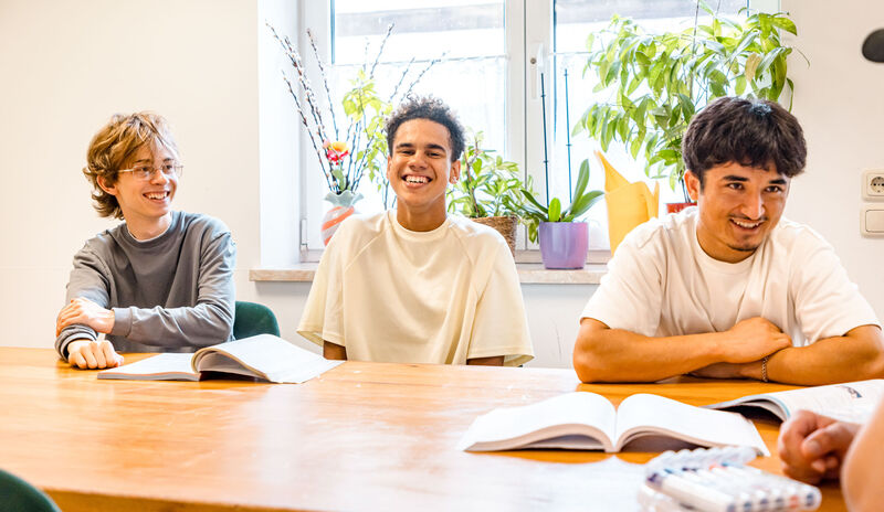 Jugendlicher sitzt lächelnd mit anderen Personen an einem Tisch in der Jugendbetreuung Jugendlicher sitzt lächelnd mit anderen Personen an einem Tisch in der Jugendbetreuung