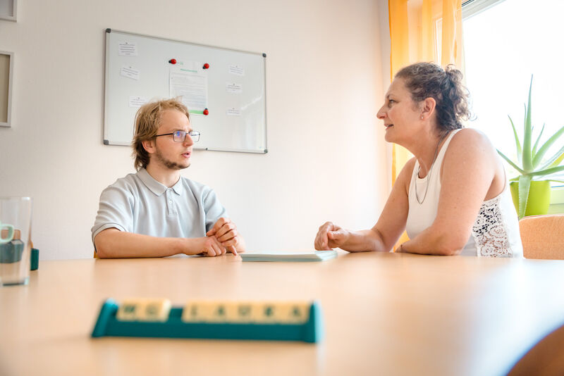 Jugendlicher und Mitarbeiterin sitzen gemeinsam an einem Tisch und führen ein Gespräch